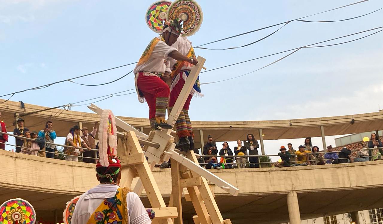 A group of circus artists in colourful costumes perform in an amphitheater doing acrobatics on tall wood structures.