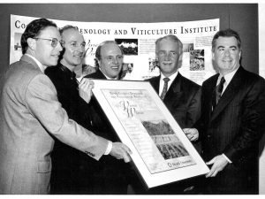 A black and white photo of five men each helping to hold up a large poster announcing the creation of the Cool Climate Oenology and Viticulture Institute.