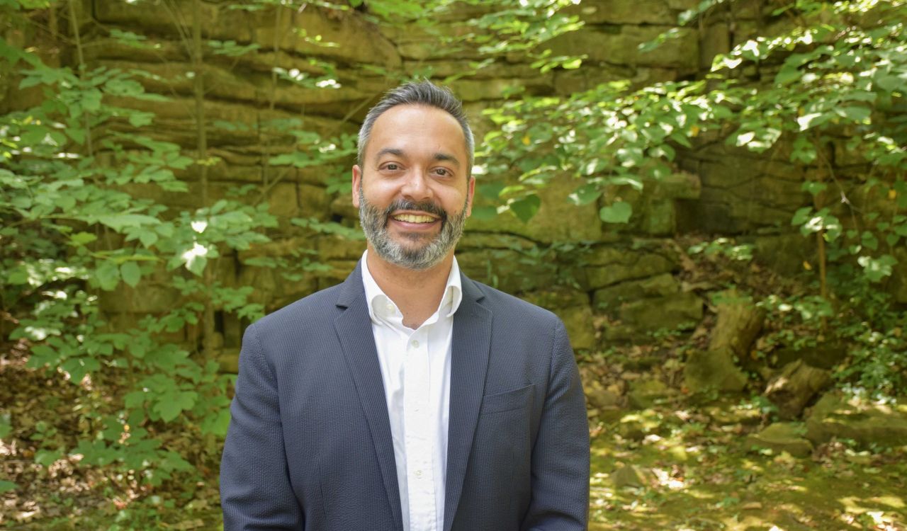 A man wearing a suit smiles at the camera standing in a green, outdoor setting.