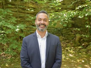 A man wearing a suit smiles at the camera standing in a green, outdoor setting.