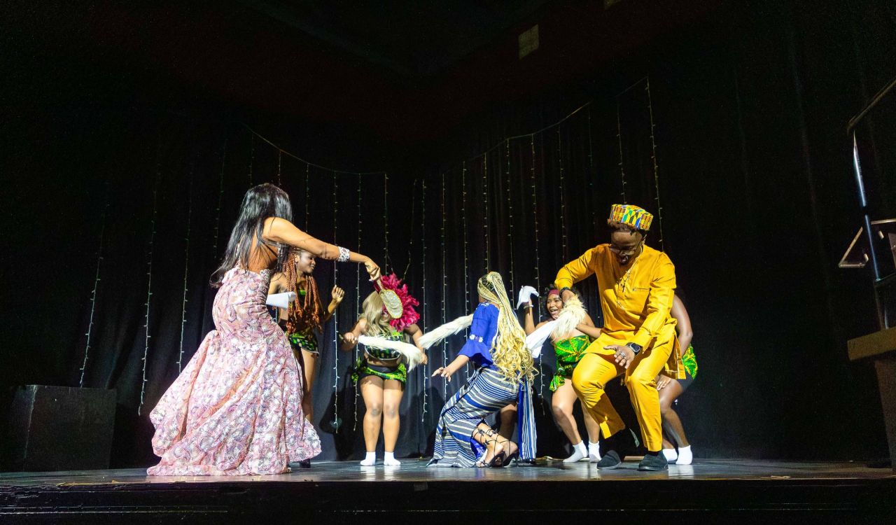 A group of young people in brightly coloured clothing dance together on a stage.