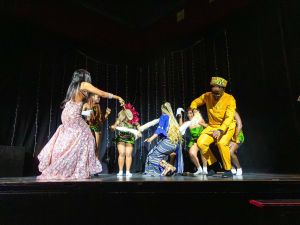 A group of young people in brightly coloured clothing dance together on a stage.