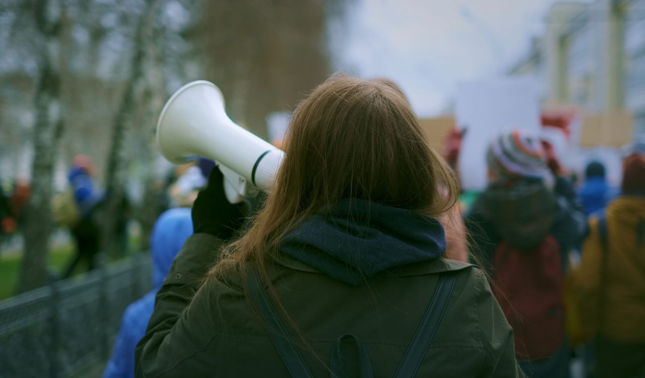 A person, seen from behind, holds up a megaphone and walks down the street during a protest.