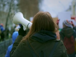 A person, seen from behind, holds up a megaphone and walks down the street during a protest.