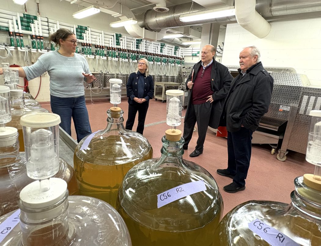 Four people stand in front of jugs of wine in a lab space.