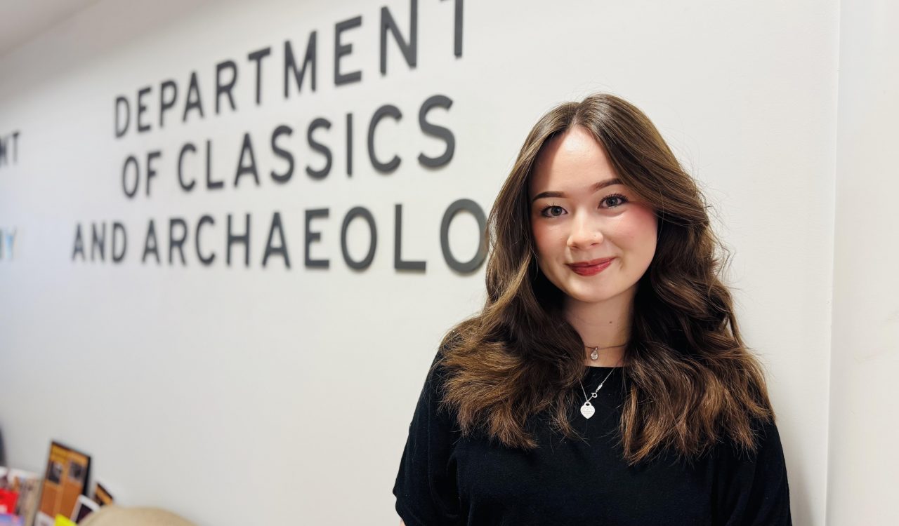 A woman smiles standing in front of a white wall with lettering that reads Department of Classics and Archaeology.