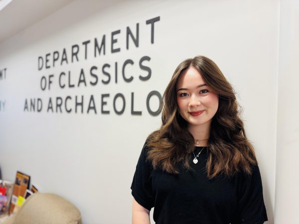A woman smiles standing in front of a white wall with lettering that reads Department of Classics and Archaeology.