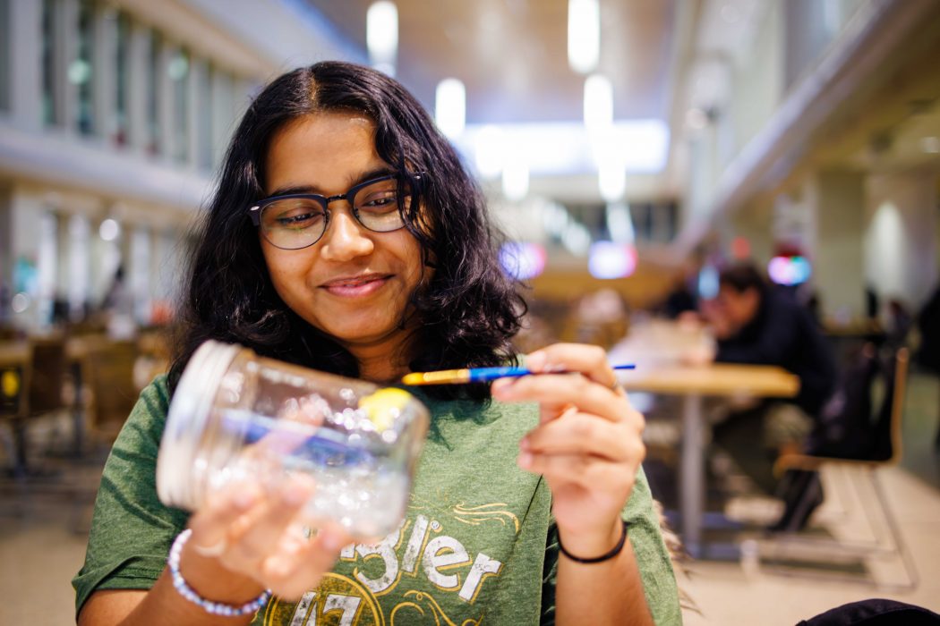 A person wearing glasses paints a yellow design onto a clear glass jar while seated indoors, with tables and other people blurred in the background.