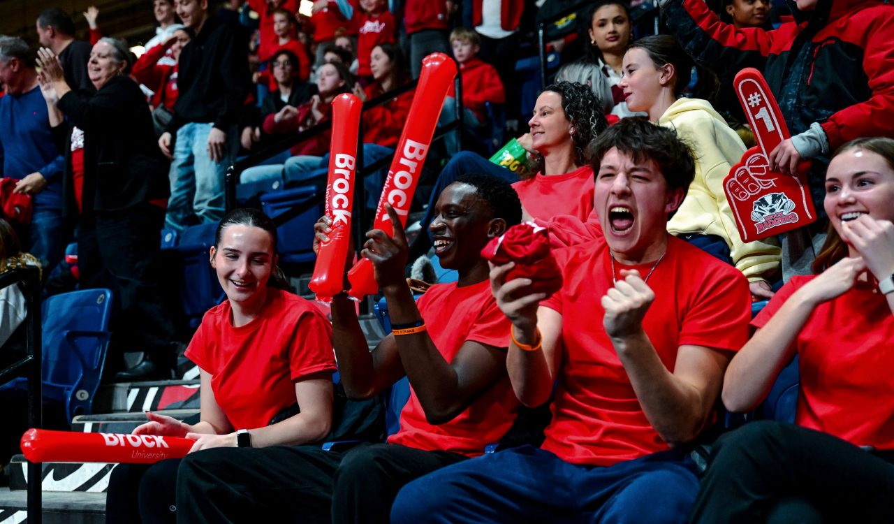 A crowd of people in red shirts cheer while sitting in stadium seats.