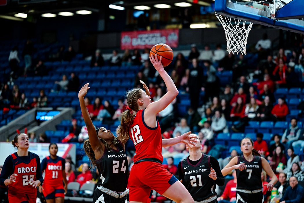A woman jumps with a basketball in her hands towards a hoop in a large arena with fans in the stands.