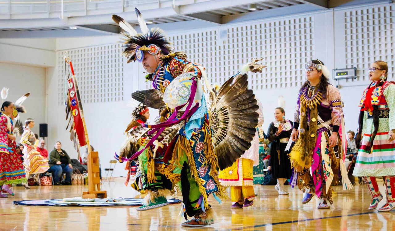 A person in Indigenous regalia dances in a gymnasium.