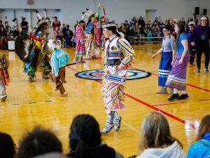 People in Indigenous regalia dance in a gymnasium as a crowd looks on.