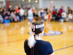 A woman in Indigenous regalia looks out over a gymnasium filled with people.