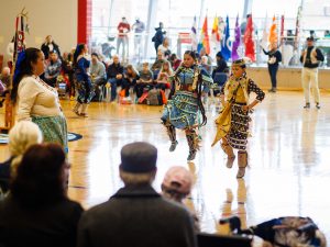 Two women in Indigenous regalia dance in a gymnasium of people.
