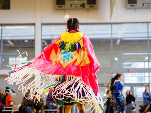The back of a person wearing brightly coloured Indigenous regalia with fringe flying through the air as they dance.