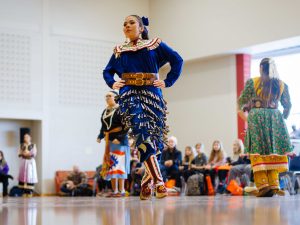 A woman in Indigenous regalia dances in a gymnasium.