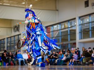 A person in Indigenous regalia dances in a gymnasium.