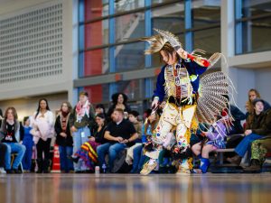 A person in Indigenous regalia dances in a gymnasium.