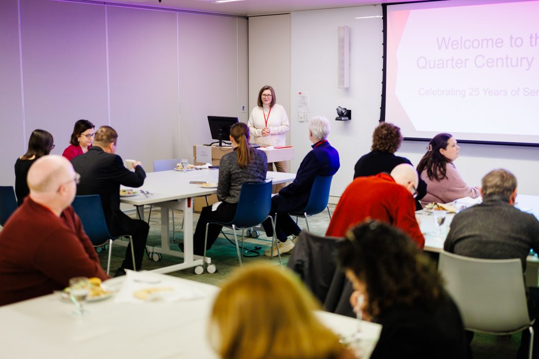 A woman stands at the front of a room speaking to guests at a lunch event.