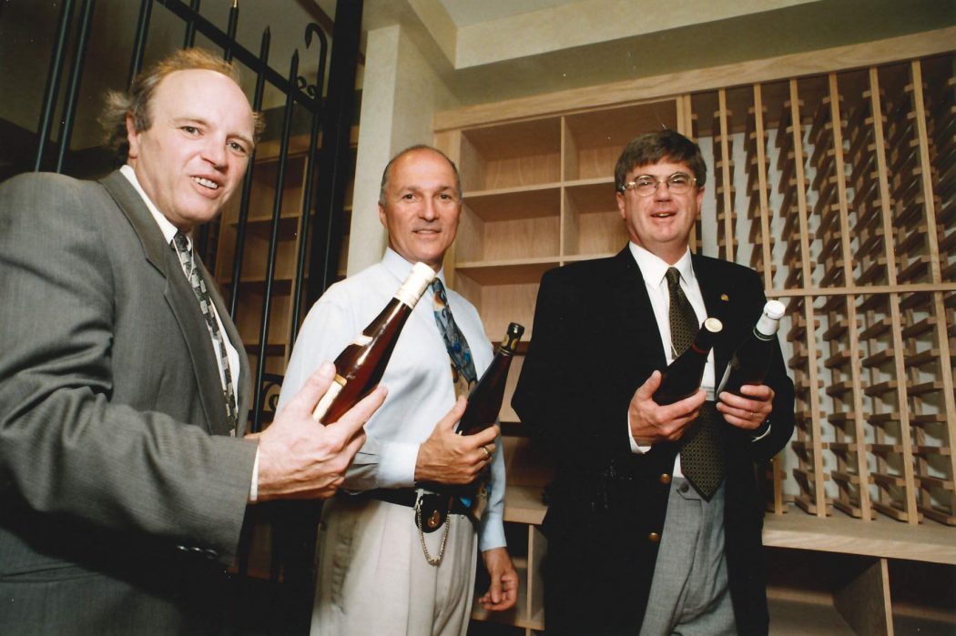 Three men pose for a picture with wine bottles in their hands.