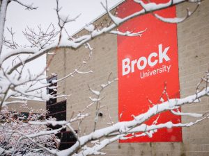 Snow-covered tree branches frame a red Brock University sign.