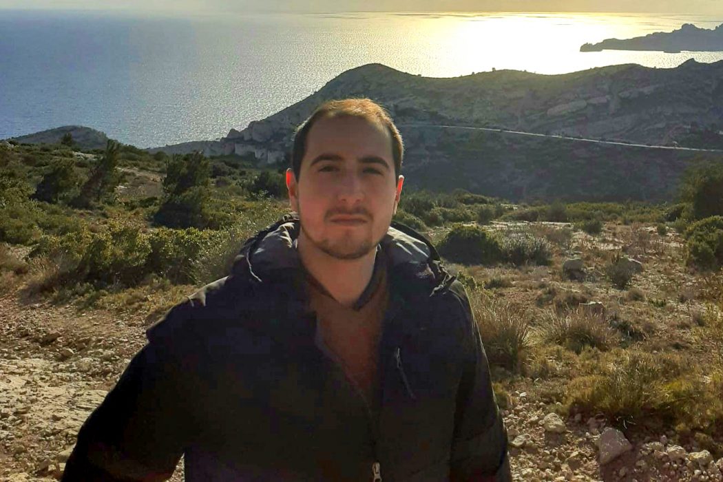 A man stands outside on a hill with a lake or ocean in the background.