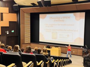 A woman stands in an auditorium speaking to an audience.