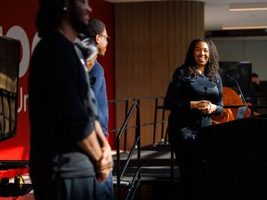 A smiling woman looks at a group of people while standing behind a podium on stage.