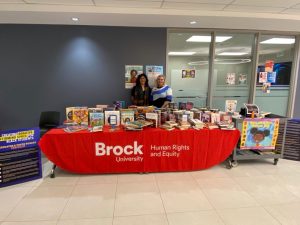 Two people stand behind a table covered in books in a hallway.