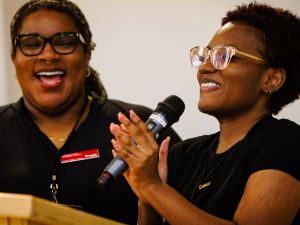 A woman holds a microphone and smiles at an unseen audience. Beside her is another woman.