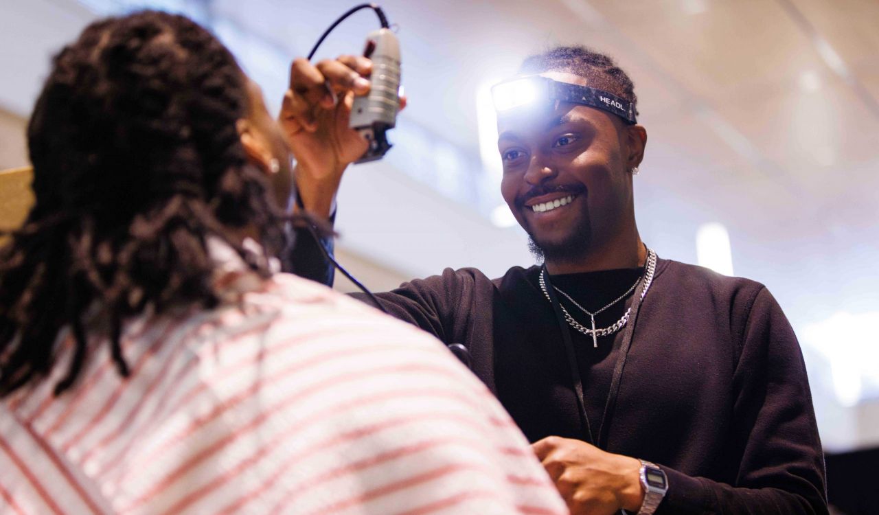 A man wearing a headlamp uses an electric hair trimmer to style someone's hair.