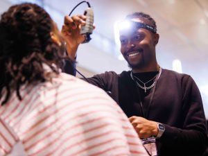 A man wearing a headlamp uses an electric hair trimmer to style someone's hair.