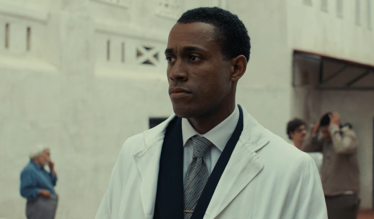A man in a white lab coat stands in a white room.