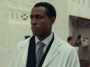 A man in a white lab coat stands in a white room.