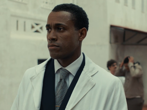 A man in a white lab coat stands in a white room.