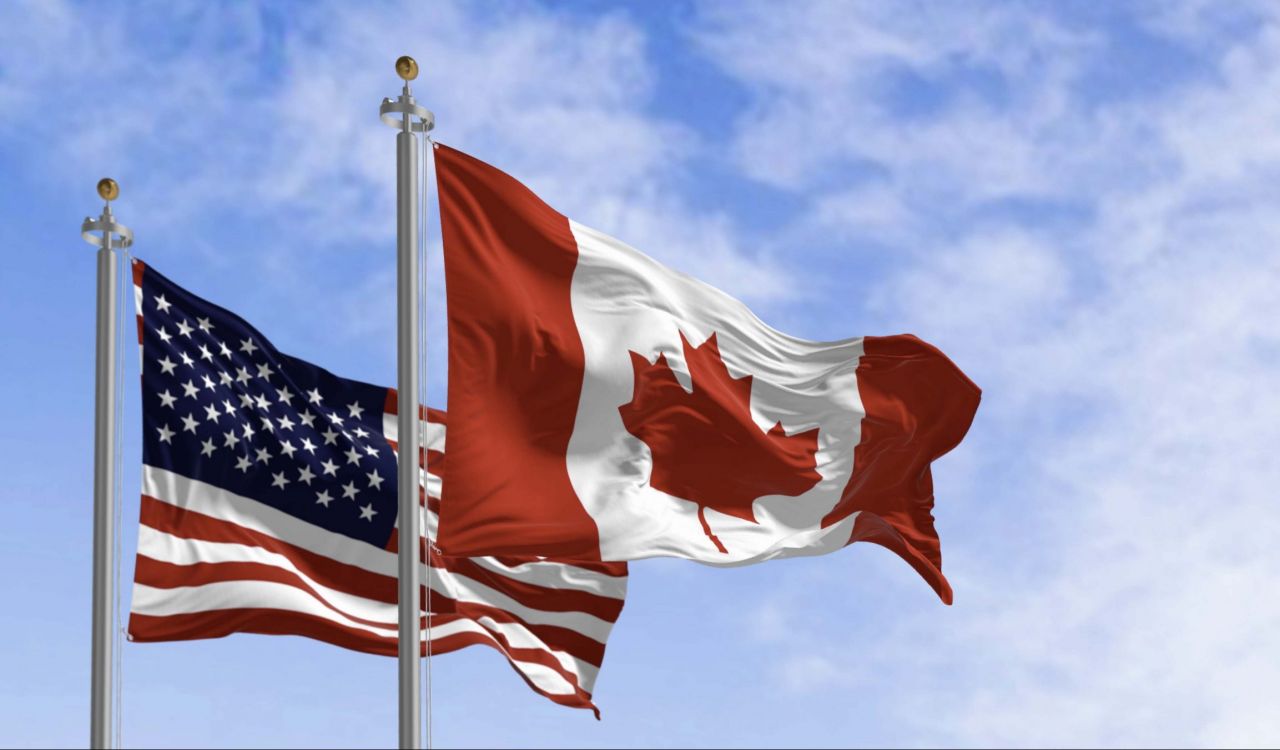 The American and Canadian flags fly against a blue sky.