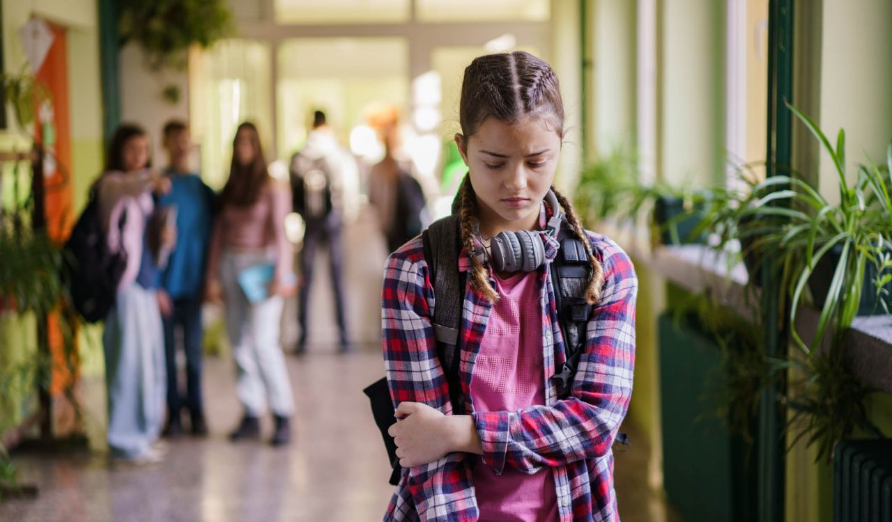 A teenager walks down the hall looking sad while other teenagers point and laugh behind them.