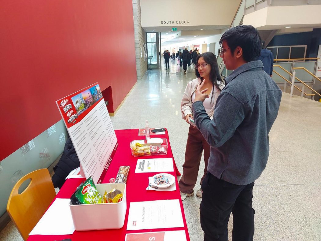 Two university students stand beside a table in a hallway having a conversation.