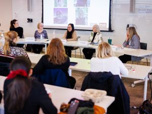 A wide view of a panel discussion shows five panelists seated at a long table speaking to an audience in a classroom setting. Name placards are visible, and a presentation slide is projected behind them.