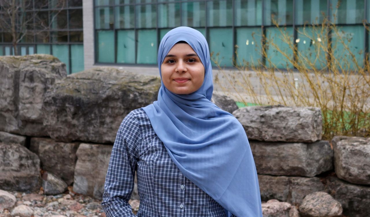 A young woman wearing a hijab poses for a photo outside a building.