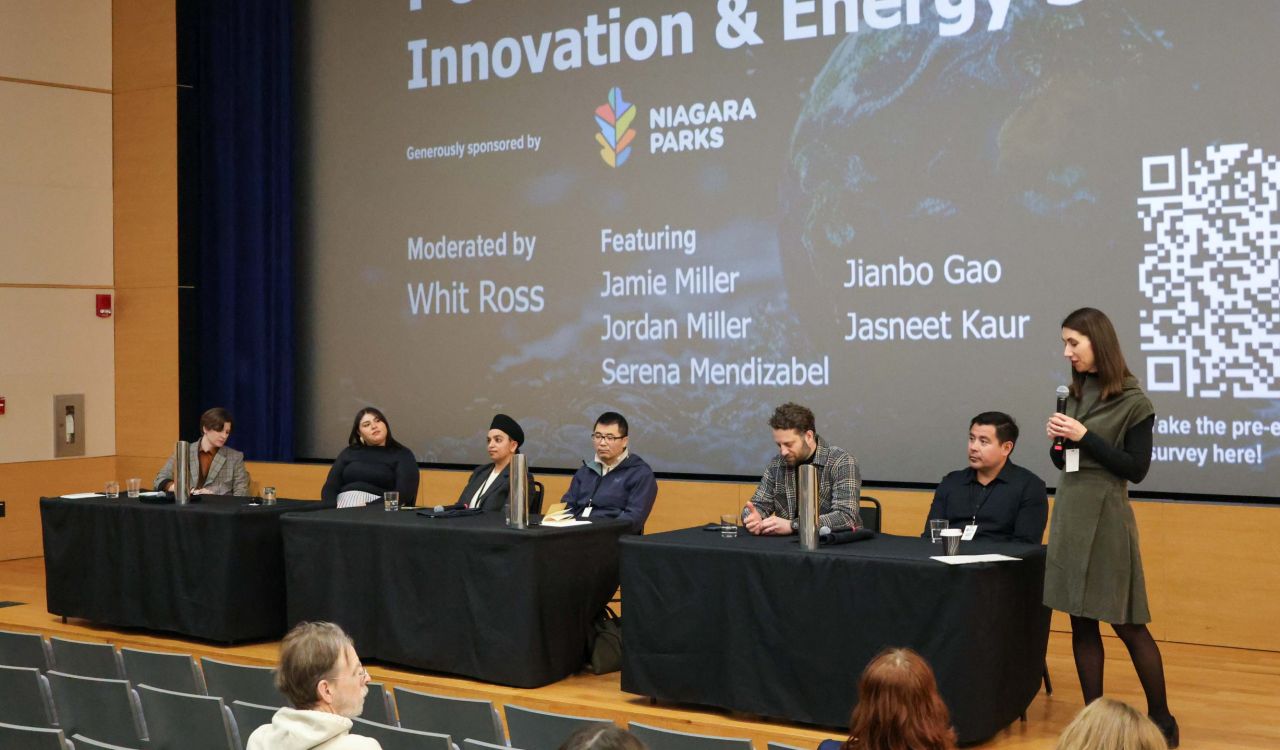 A group of people sit at a table on stage during a panel while a woman holding a microphone stands nearby.