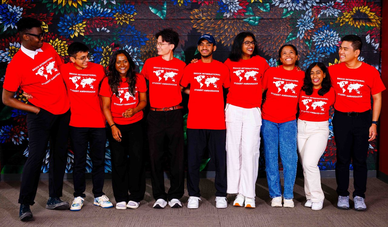 A group of university students in red shirts pose in front of a floral mural.