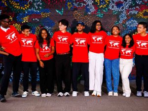 A group of university students in red shirts pose in front of a floral mural.