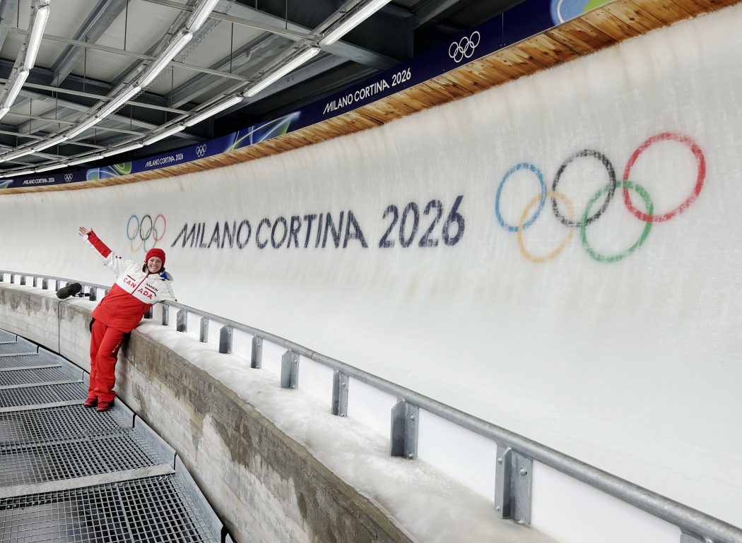 A woman in Team Canada gear stands at an ice bobsleigh track with the Milano Cortina logo and Olympic rings displayed on the ice.