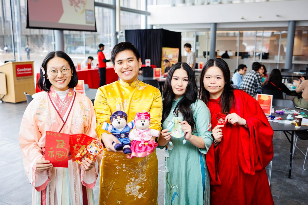 Four people stand side by side indoors at a Lunar New Year event, smiling at the camera while wearing colorful traditional outfits.