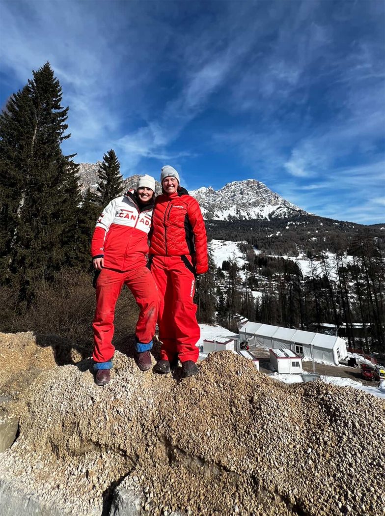 Two women in Team Canada clothing stand on mountainous terrain with snow-peaked mountains in the background.