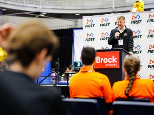 A man speaks to an audience from a podium during a high school robotics competition.