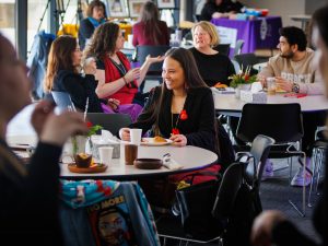 A group of attendees sit at round tables in a bright gathering space, talking and sharing refreshments during a REDress Day event.