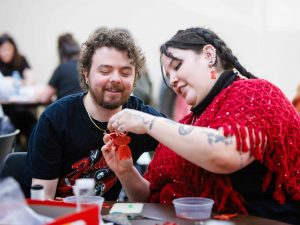 Two participants sit together crafting a small red dress ornament at a table with beads and materials.