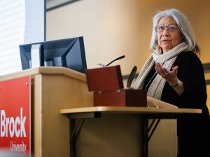 An older woman speaks at a podium with a ‘Brock University’ sign, addressing the audience during a REDress Day gathering.